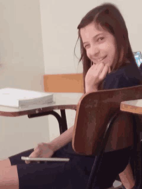 a girl sits at a desk with her hand on her chin smiling
