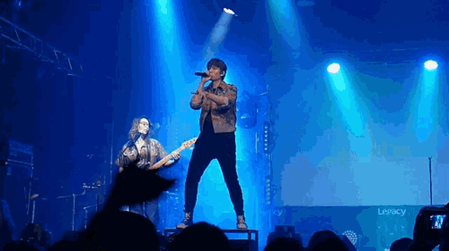 a man singing into a microphone on a stage in front of a legacy logo