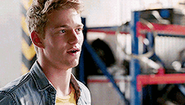 a young man wearing a denim jacket and a yellow shirt is standing in a warehouse .