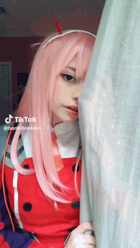 a girl with pink hair and horns is peeking out from behind a curtain in a room .