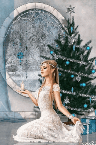 a woman in a white dress is kneeling in front of a christmas tree and holding a candle
