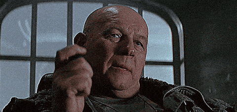 a bald man is sitting in front of a window and pointing his finger at the camera .