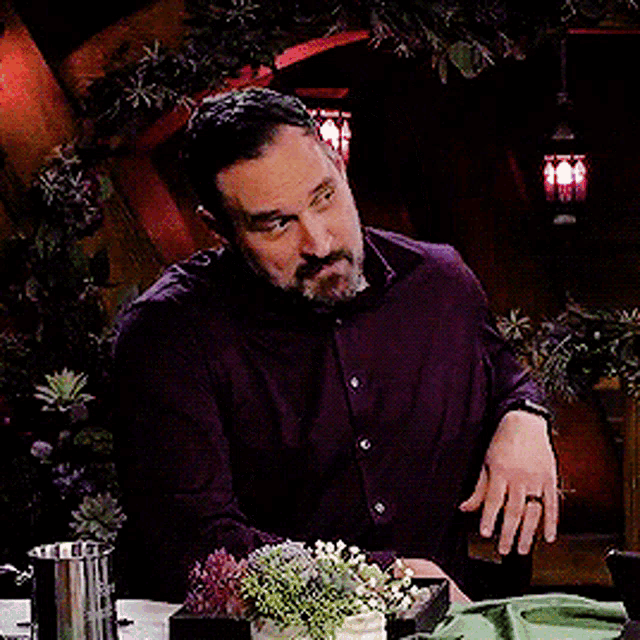 a man with a beard is sitting at a table with a potted plant