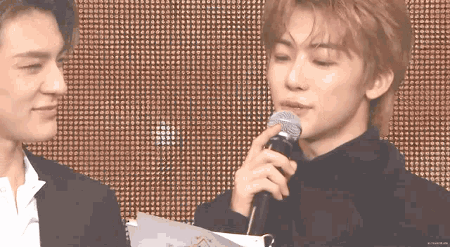 two men are standing next to each other and one is holding a microphone and the other is holding a piece of paper .