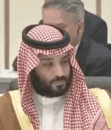 a man with a beard wearing a keffiyeh and a robe is sitting in a chair .