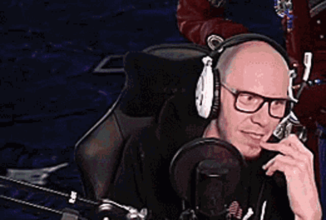 a bald man wearing headphones and glasses sitting in front of a microphone