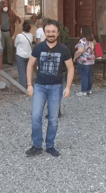 a man in a black t-shirt and blue jeans is standing on a gravel road .