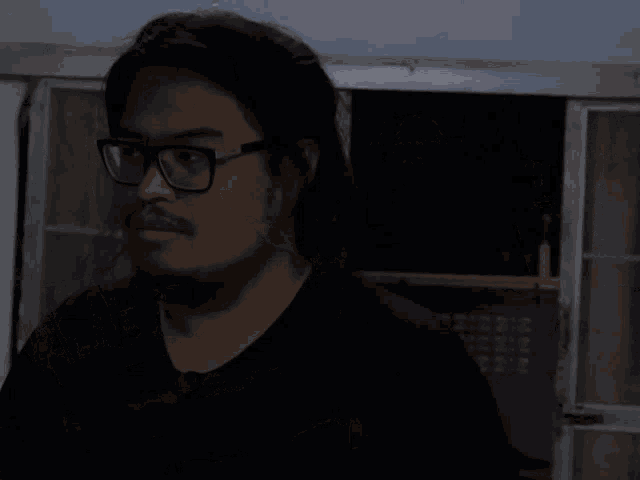 a man wearing glasses and a black shirt is smiling in front of a window with a foreign language caption