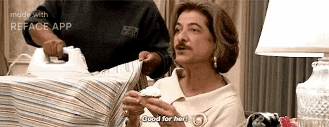 a woman is holding a cupcake and saying `` good for her '' while a man irons clothes in the background .