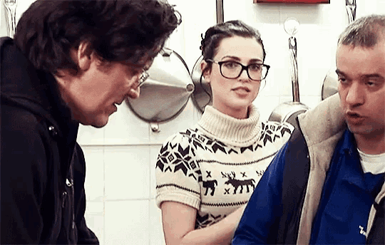 a woman wearing glasses talks to two men in a kitchen .