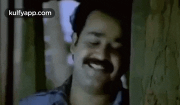 a man with a mustache is smiling while leaning against a wall in a room .