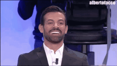 a man with a beard wearing a suit and white shirt is smiling in front of a camera .