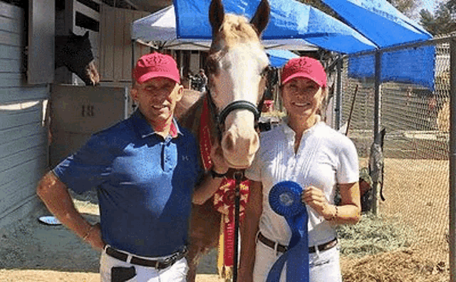 a man and a woman are standing next to a horse and holding a blue ribbon .