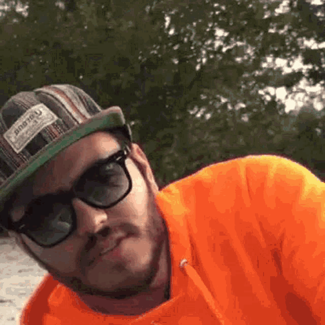 a man wearing sunglasses , a hat and an orange hoodie is making a funny face .