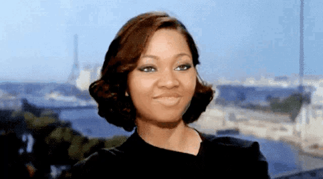 a woman in a black dress looks at the camera with a city in the background