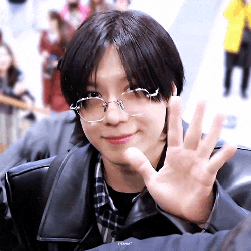 a young man wearing glasses and a black jacket is waving his hand