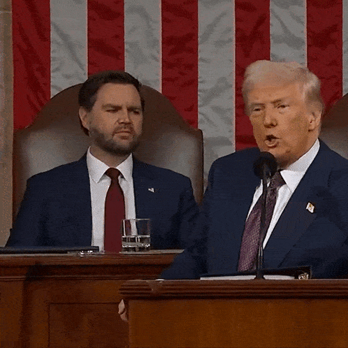 a man in a suit and tie is speaking into a microphone while another man looks on with the word lunatics behind him
