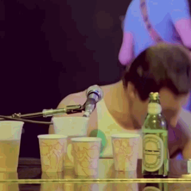 a man is drinking from a bottle while sitting at a piano with cups and a microphone
