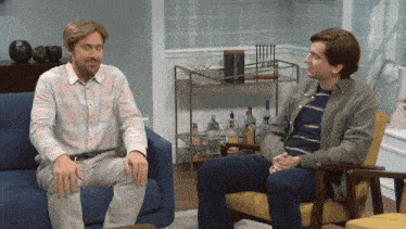 two men are sitting on a couch and one is wearing a wig