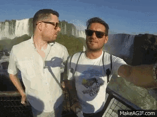 two men taking a selfie in front of a waterfall with makeagif.com in the corner
