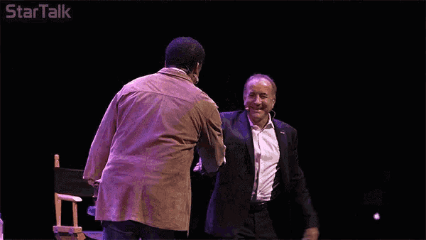 a man in a suit shakes hands with another man in a brown jacket on a stage with startalk written on the bottom