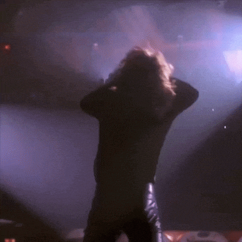 a man in a black shirt and leather pants is standing on a stage with his hands behind his head .