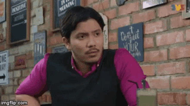 a man in a pink shirt and black vest is sitting in front of a brick wall .