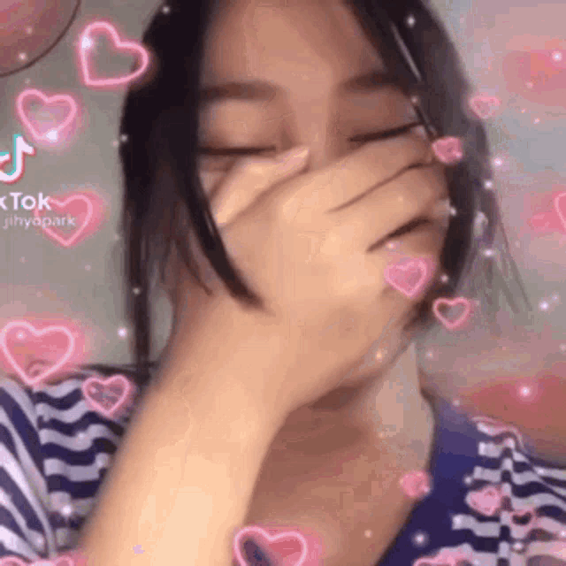 a girl is covering her face with her hand and there are pink hearts flying around her .