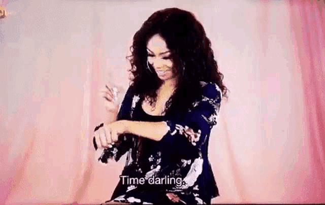 a woman with curly hair is sitting in front of a pink curtain and smiling while looking at her watch .