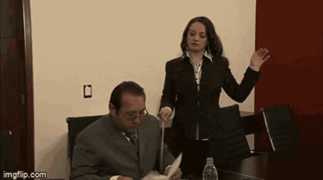 a man in a suit sits at a table while a woman stands behind him holding a piece of paper