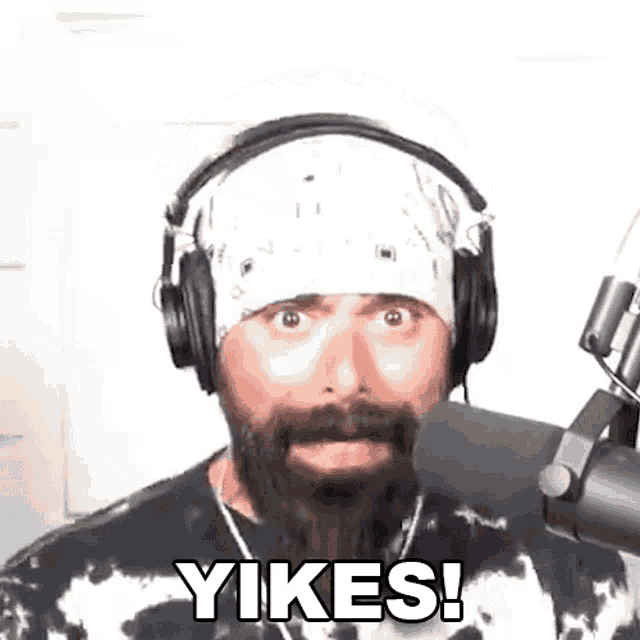 a man with a beard and headphones is standing in front of a microphone and saying yikes .