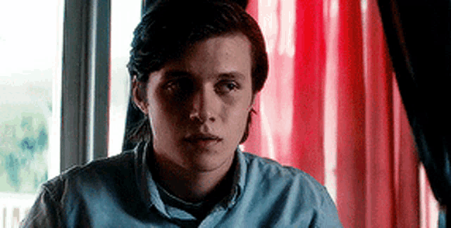 a young man in a blue shirt is sitting in front of a window with a red curtain .