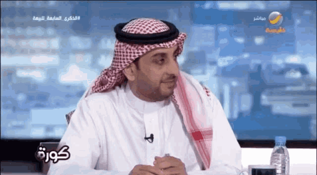 a man wearing a head scarf and a white shirt is sitting in front of a screen with arabic writing on it