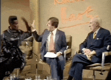 a man in a suit is talking to another man in a suit and tie while another man sits in a chair .