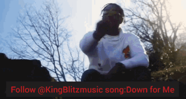 a man sits on a rock in front of trees with a follow @kingblitzmusic song down for me banner