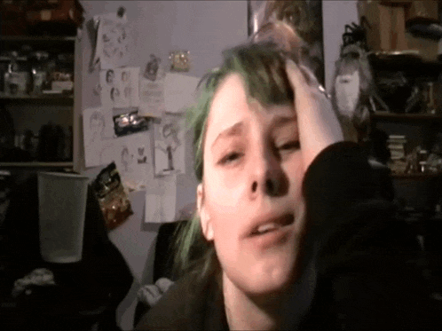 a girl with green hair holds her head in front of a wall with drawings on it