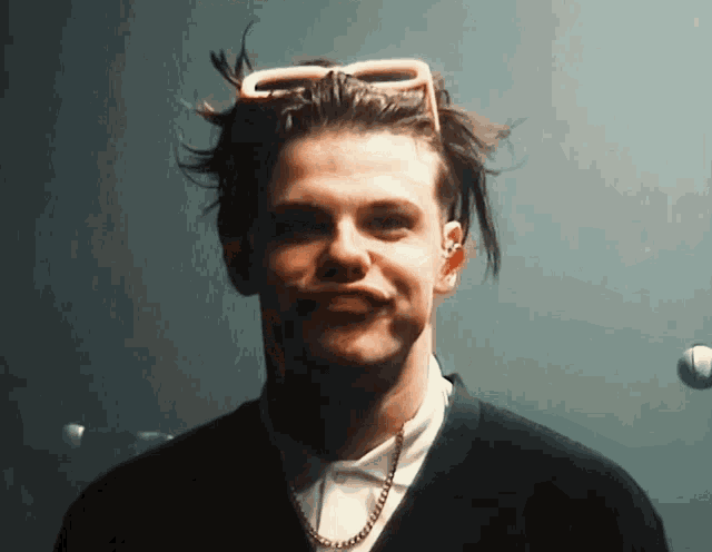 a young man wearing sunglasses and a chain around his neck looks at the camera