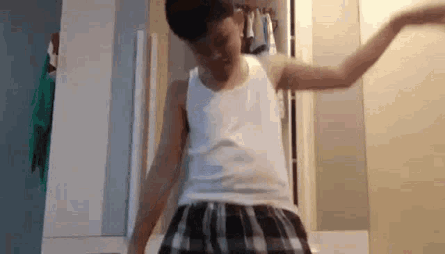 a young boy in a white tank top and plaid shorts is dancing in front of a mirror .