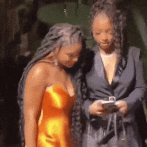 two women are standing next to each other looking at their phones . one of the women is wearing an orange dress .