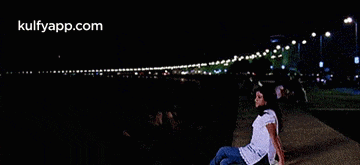 a woman is sitting on a sidewalk at night looking out over a body of water .