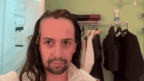 a man with long hair and a beard is standing in front of a closet with clothes hanging on it .