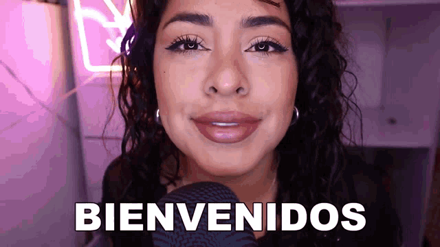 a woman wearing headphones says bienvenidos in front of a pink neon sign