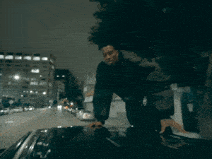 a man in a black jacket stands on the hood of his car
