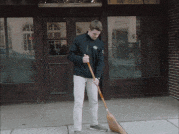 a man wearing a black jacket with a s on it is sweeping the sidewalk