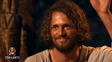 a man with curly hair and a beard is smiling and waving his hand with koh-lanta in the background