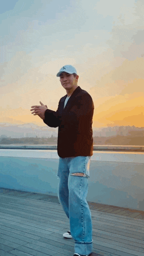 a man wearing a hat and jeans is standing on a rooftop at sunset .