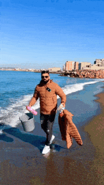 a man in a brown jacket is walking on the beach carrying a bucket and a jacket