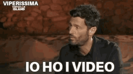 a man with a beard is sitting in front of a stone wall with the words `` io ho i video '' .