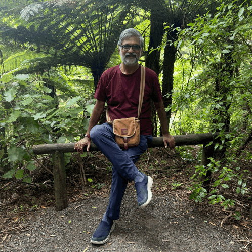 a man sits on a wooden railing in the woods with his legs crossed