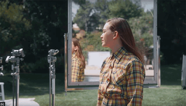 a woman in a plaid shirt stands in front of a mirror with the letter k in the corner